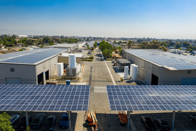 Aerial view of a commercial facility with rooftop and carport solar panel installations, where the carport both shelters vehicles and generates renewable energy. Two large warehouse buildings are covered in sleek photovoltaic panels, surrounded by residential neighborhoods.